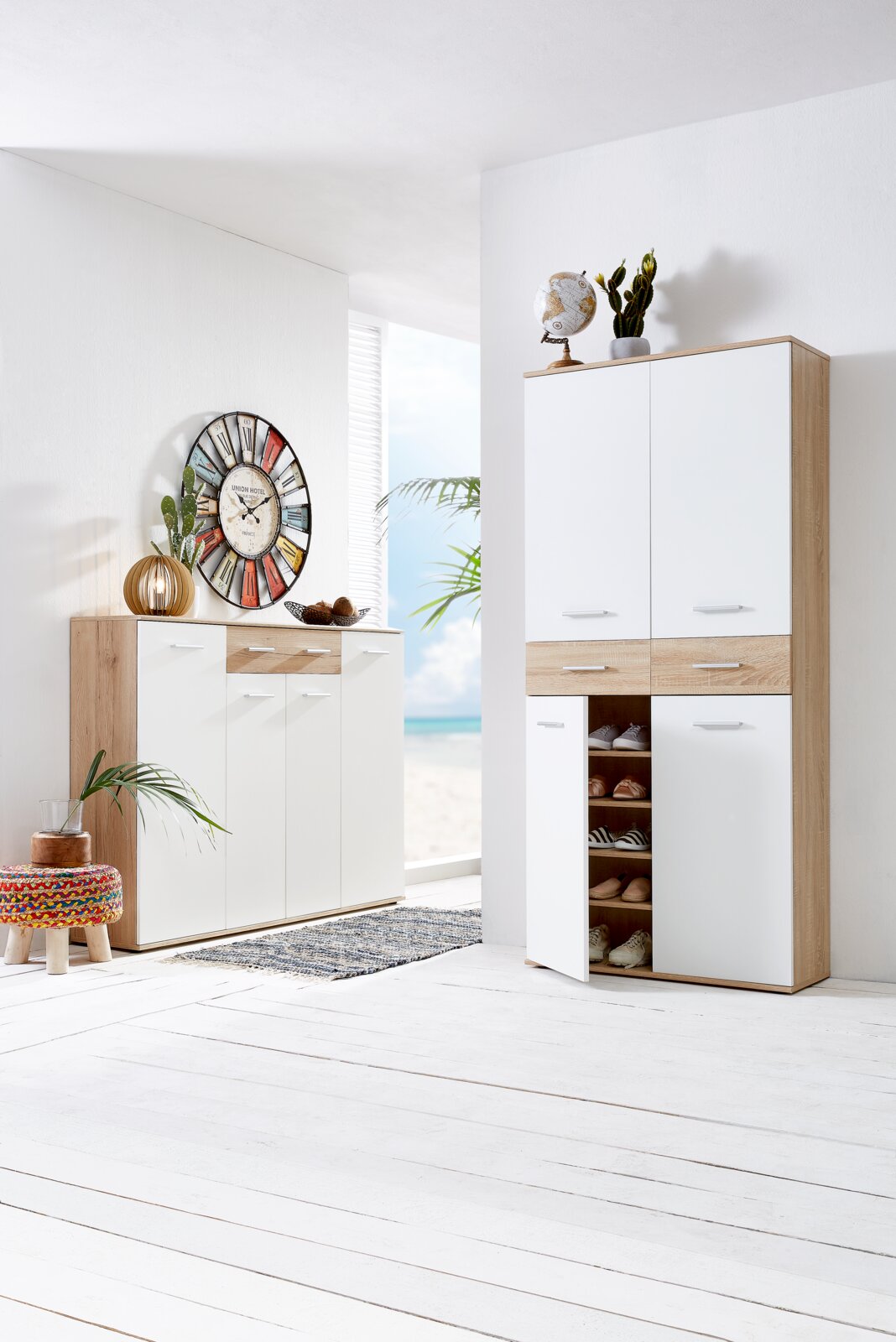 XORA XL-schoenenkast FAKIR Moderne XL-schoenenkast in lichte houtlook met witte deuren, gedeeltelijk open, in een lichte kamer met uitzicht op het strand.