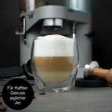 Nahaufnahme eines Latte Macchiato in einem doppelwandigen Glas vor einer Kaffeemaschine, mit Kaffeebohnen und Tamper im Vordergrund.