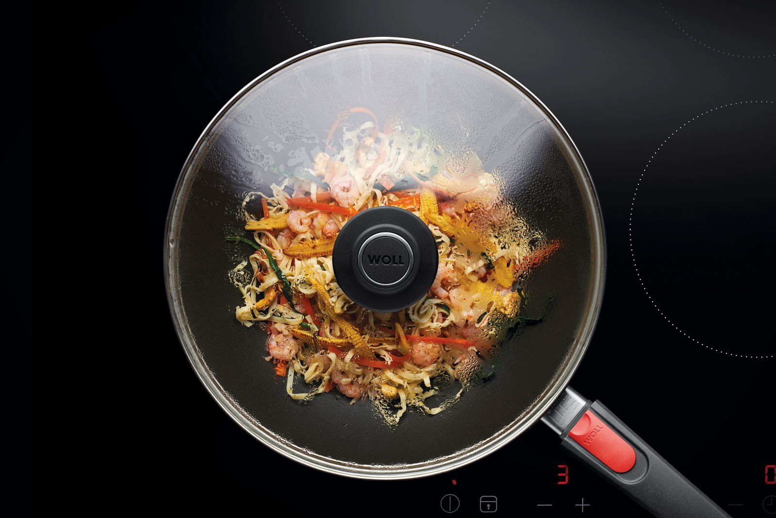 Guss-Schwenkpfanne mit Glasdeckel, gefüllt mit buntem Gemüse und Garnelen, von oben fotografiert auf einem Herd.