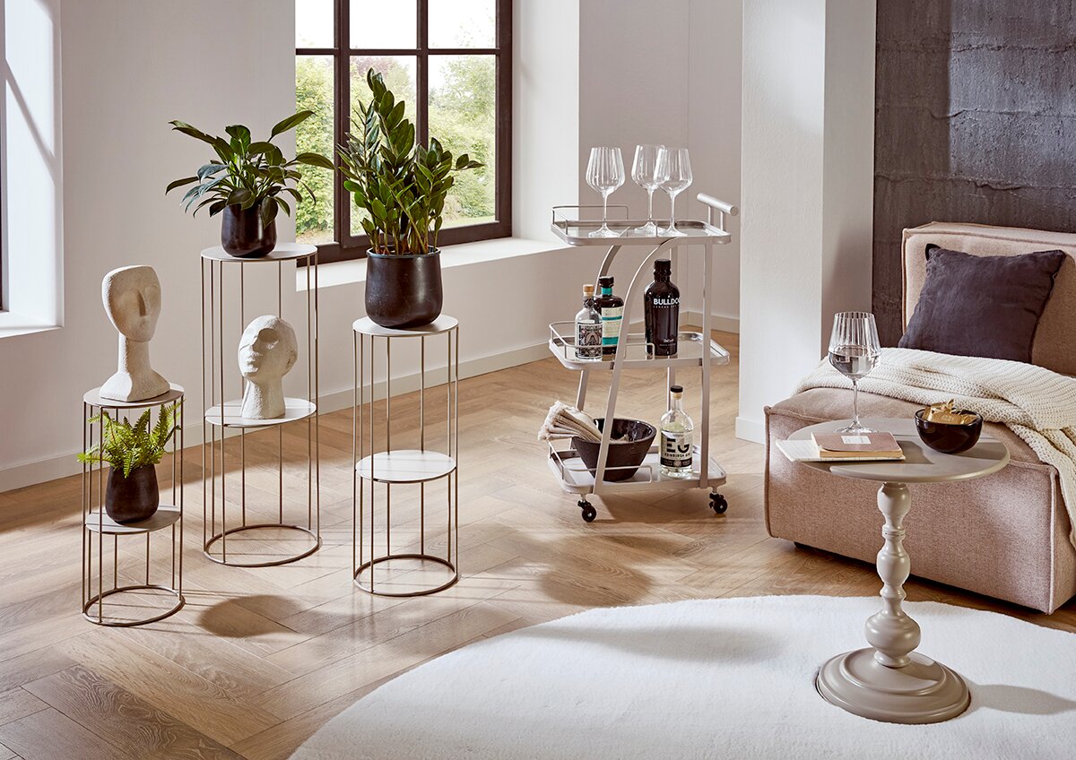 Zurbrüggen Blumensäule Helles Wohnzimmer-Setting mit weißen Metall-Pflanzenständern, beigem Servierwagen und kleinem Beistelltisch neben Sofa, fotografiert in leichter Seitenansicht aus Augenhöhe.