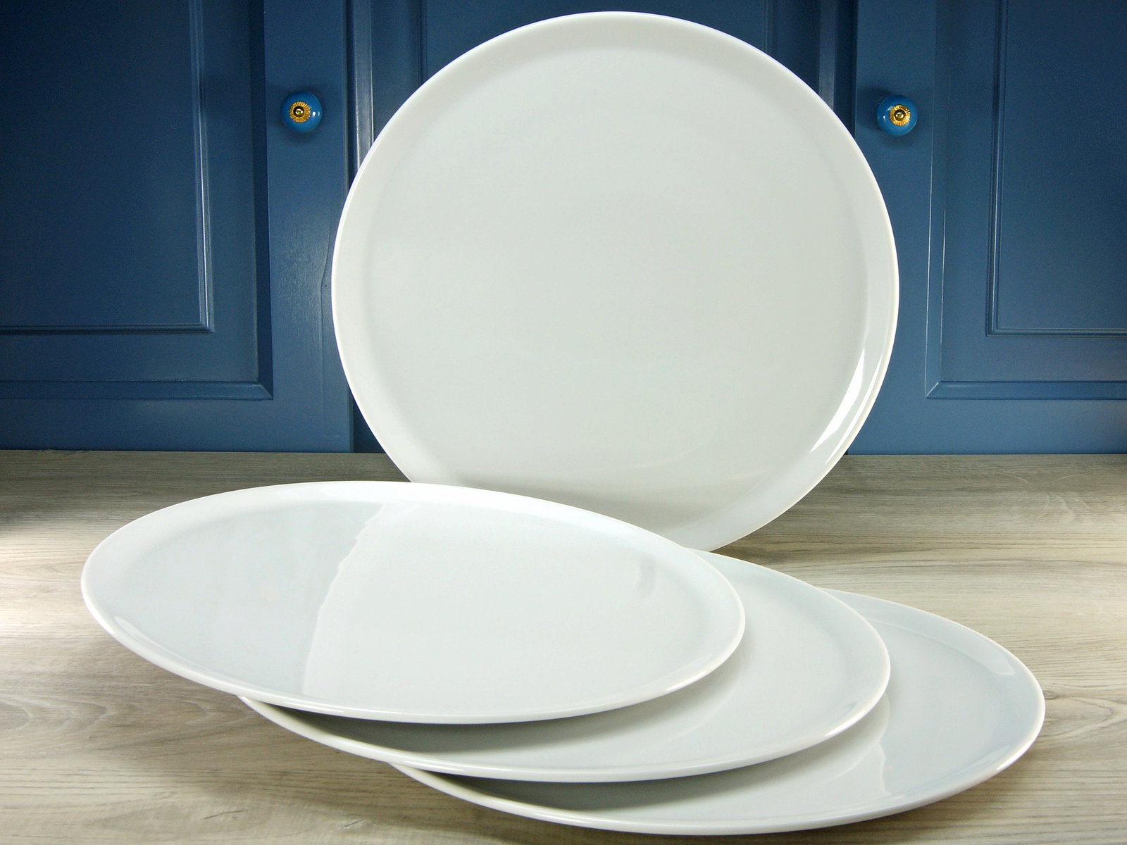 Vier weiße Pizzateller, einer aufrecht stehend und drei flach liegend, vor einem blauen Hintergrund.