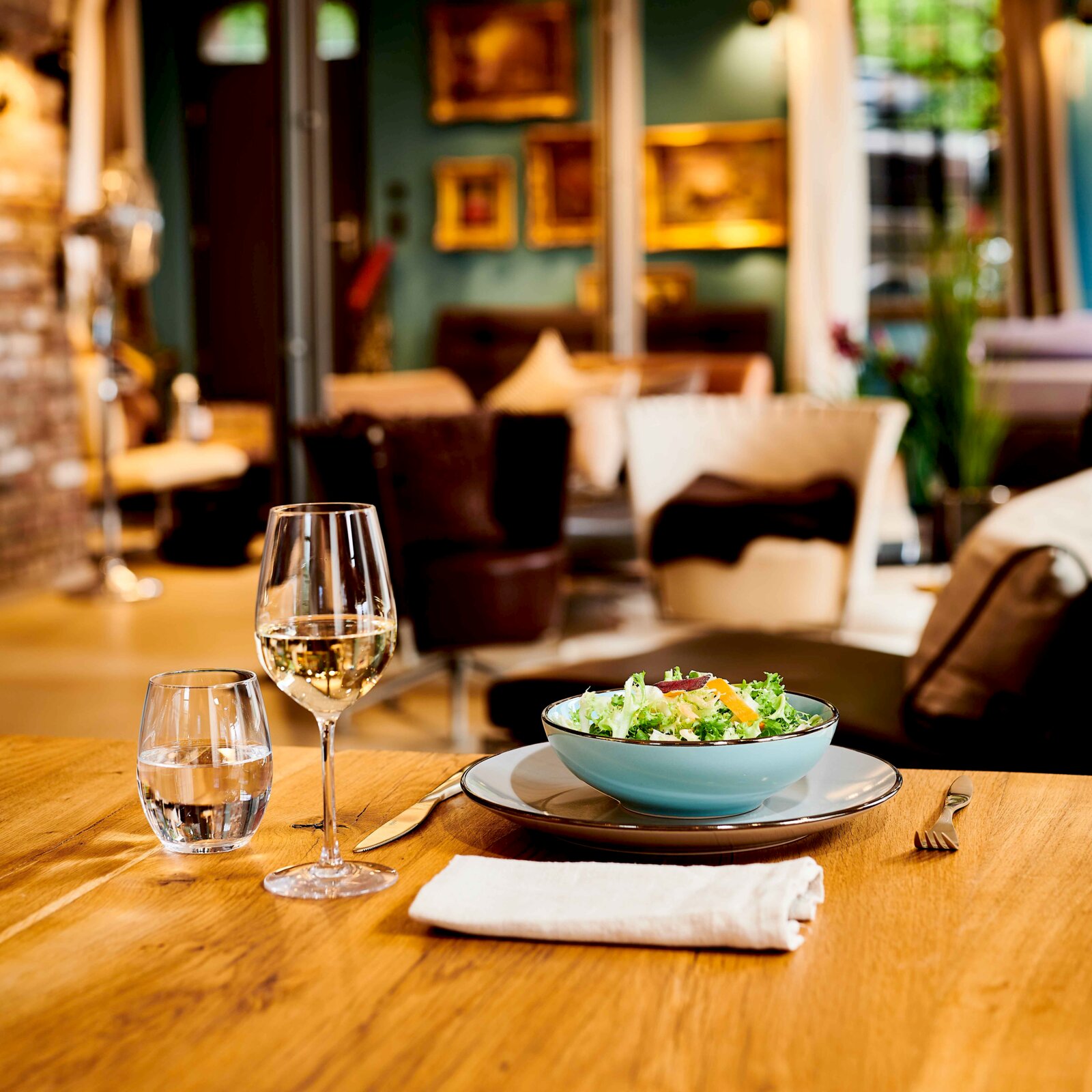 Nahaufnahme aus Tischperspektive: Salatschüssel auf Teller mit Besteck, daneben Wasser- und Weinglas auf Holztisch in gemütlichem Restaurant, Hintergrund unscharf