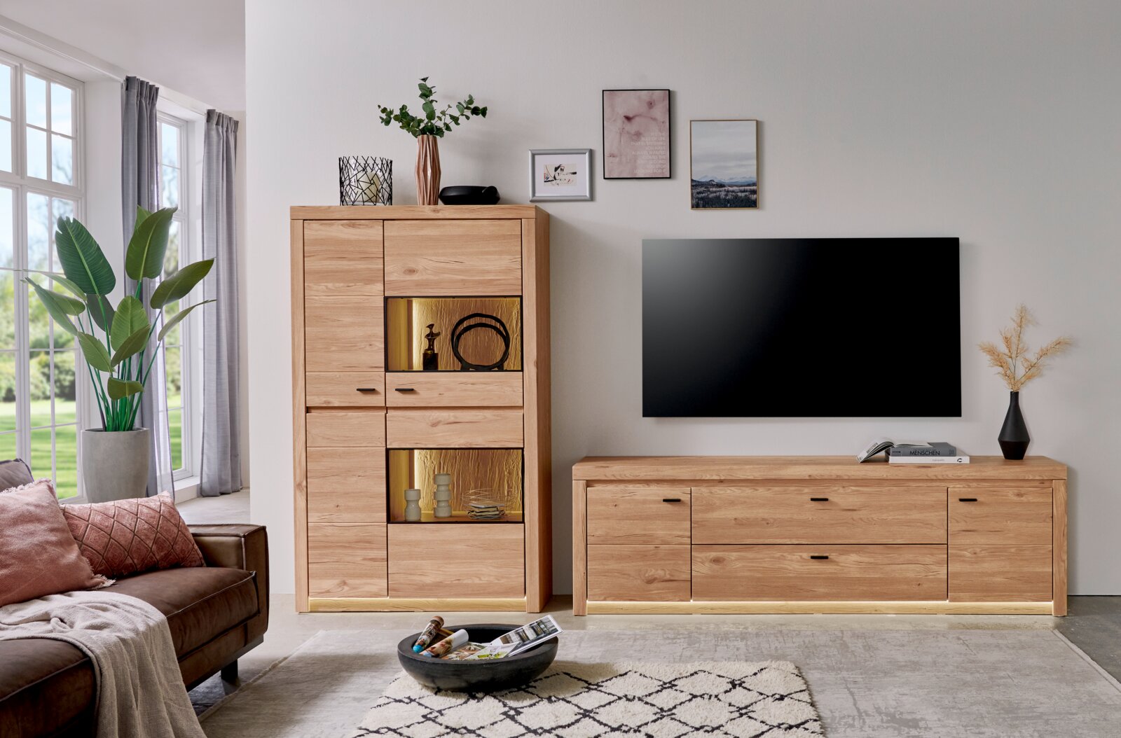 Modernes Wohnzimmer-Set aus hellem Holz mit Vitrine/Highboard mit beleuchteten offenen Fächern und passendem TV-Lowboard unter einem wandmontierten Fernseher, frontal aus leichter Seitenperspektive aufgenommen.