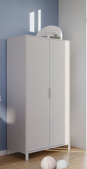 Moderner weißer Kleiderschrank mit zwei Türen, seitliche Perspektive, auf hellem Holzboden vor blauer Wand, mit Plüschtier obenauf.