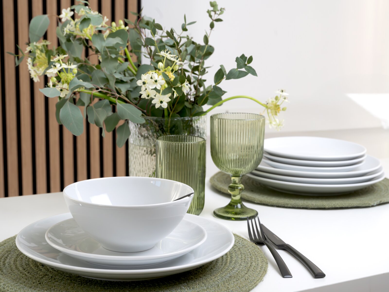 Schlichtes weißes Geschirr-Set mit Schüssel auf Teller, gestapelten Tellern und grünen geriffelten Trinkgläsern neben einer Vase mit Eukalyptus und weißen Blüten, in leichter schräger Seitenansicht auf einem gedeckten Tisch.