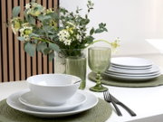 Schlichtes weißes Geschirr-Set mit Schüssel auf Teller, gestapelten Tellern und grünen geriffelten Trinkgläsern neben einer Vase mit Eukalyptus und weißen Blüten, in leichter schräger Seitenansicht auf einem gedeckten Tisch.