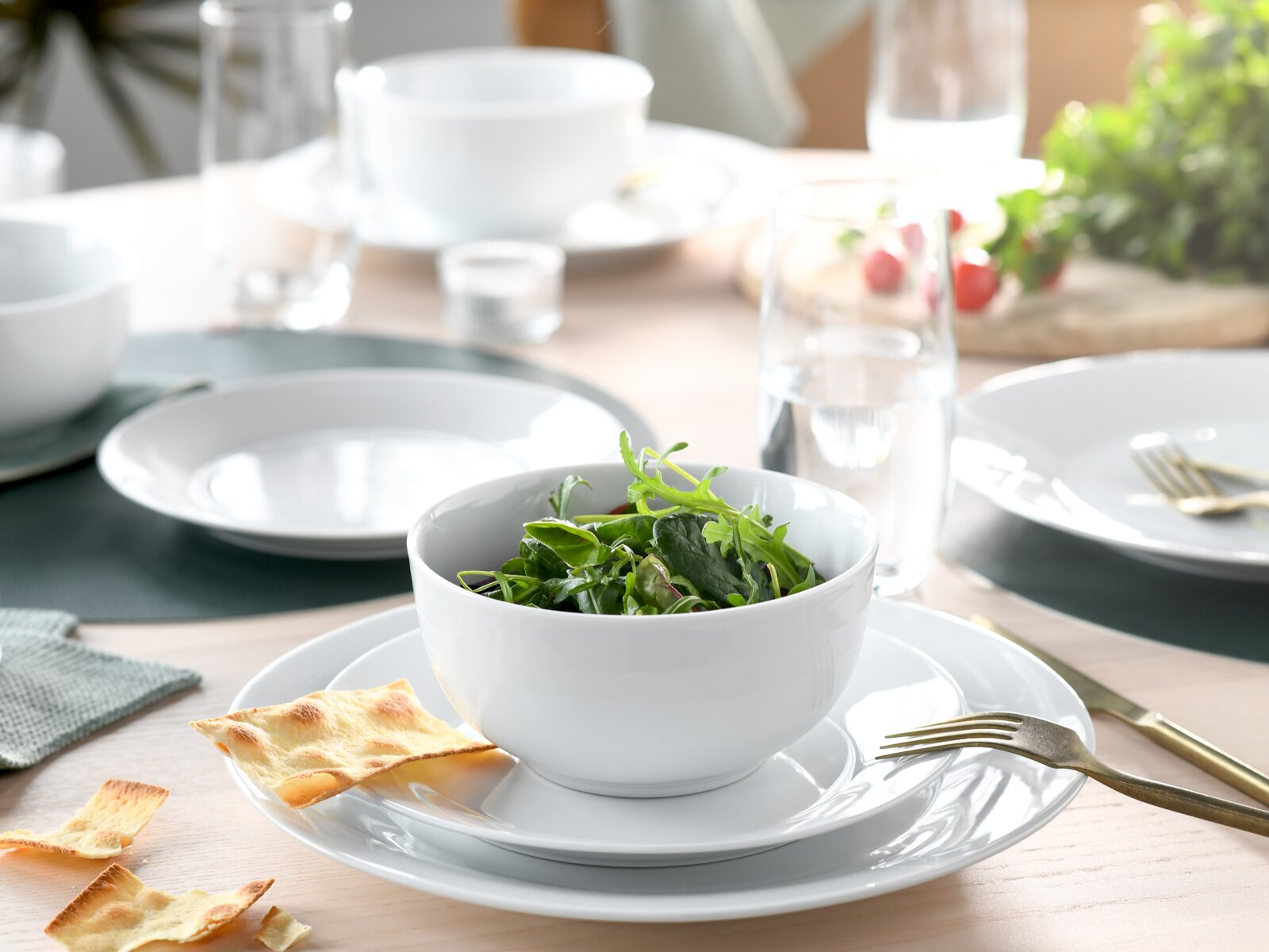 Weißes Geschirr-Set mit Schüssel und Tellern auf gedecktem Tisch, vorn eine Schüssel mit grünem Salat, seitliche Perspektive in leicht erhöhter Ansicht