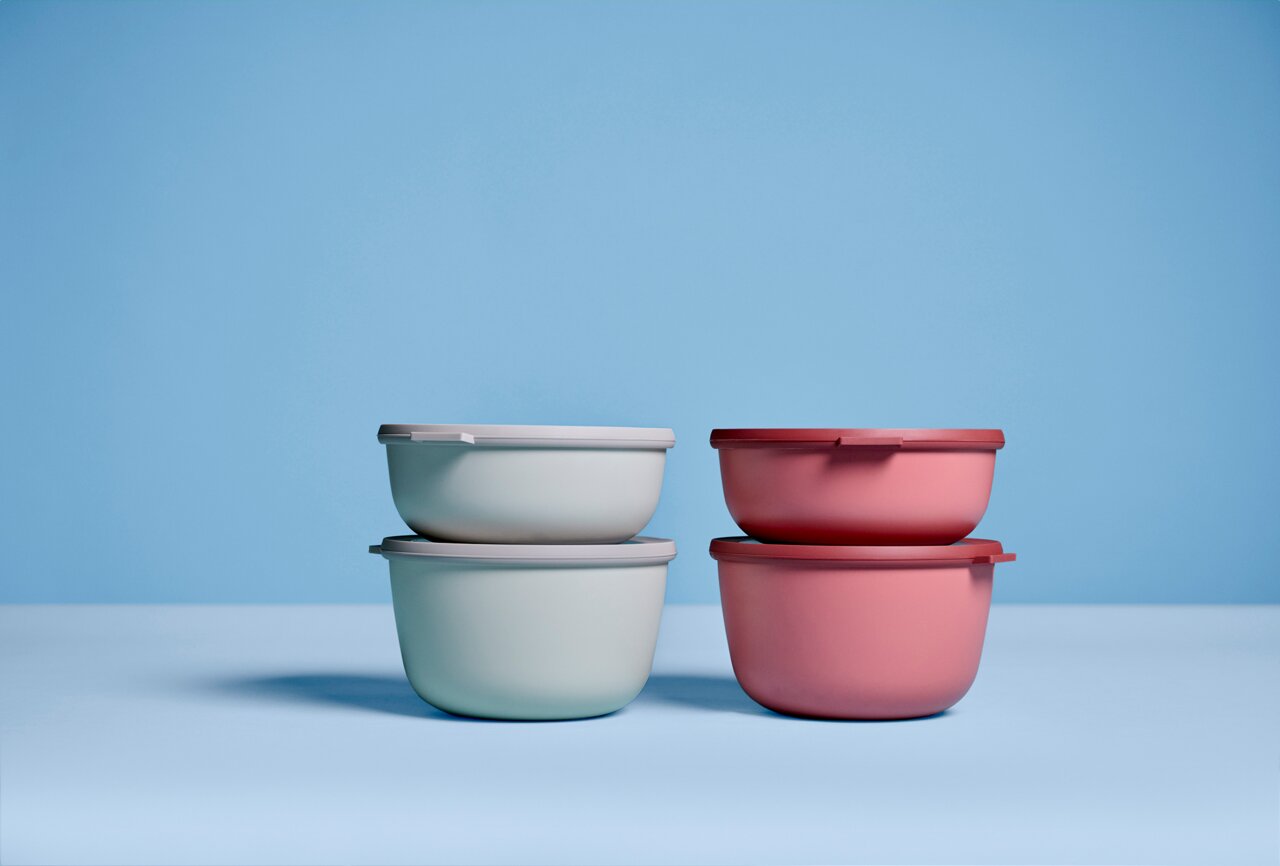 Vier stapelbare Schüsseln in Pastellfarben, zwei in Vivid Mauve und zwei in einem hellen Grünton, auf einer blauen Oberfläche vor einem blauen Hintergrund, seitliche Perspektive.