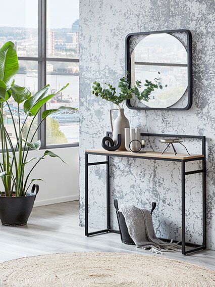 Frontale Innenraumaufnahme eines modernen Flurs: schmaler Konsolentisch mit schwarzem Metallrahmen und Holzplatte vor strukturierter grauer Wand, darüber quadratischer Wandspiegel; dekoriert mit Vase, Pflanzenzweigen und Accessoires, links großes Fenster 