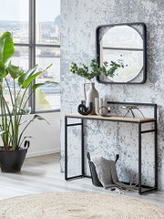 Frontale Innenraumaufnahme eines modernen Flurs: schmaler Konsolentisch mit schwarzem Metallrahmen und Holzplatte vor strukturierter grauer Wand, darüber quadratischer Wandspiegel; dekoriert mit Vase, Pflanzenzweigen und Accessoires, links großes Fenster 