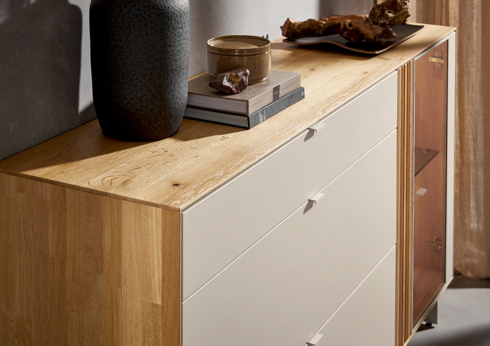 Modernes Sideboard aus hellem Holz mit weißen Fronten und Glastür, dekoriert mit Vase und Büchern, in schräger Perspektive von vorne links