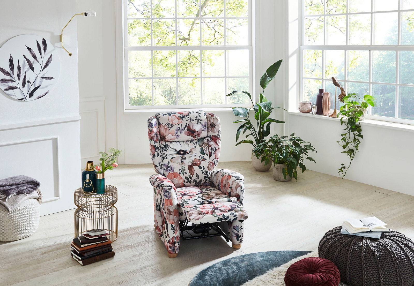 Blumiger Fernsehsessel in einem hellen Wohnzimmer mit großen Fenstern, umgeben von Pflanzen, aus der Vorderansicht.