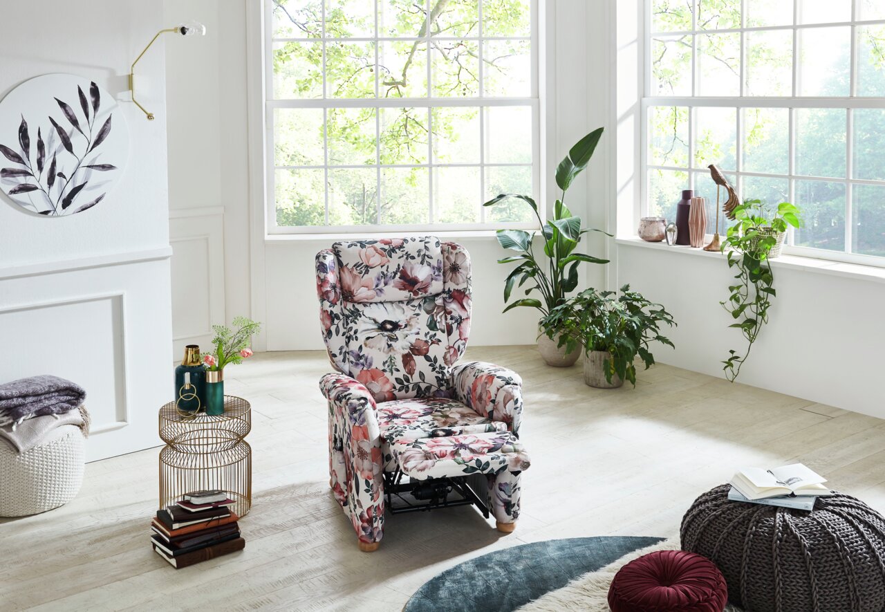 Een bloemrijke tv-fauteuil in een lichte woonkamer met grote ramen, omringd door planten, gezien van voren.