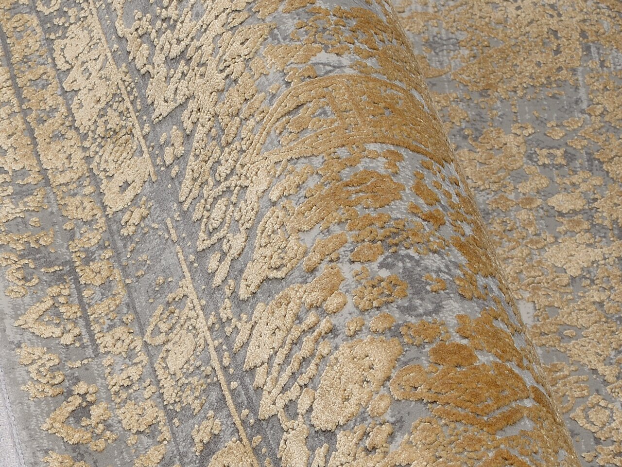 Nahaufnahme eines Designer Teppichs mit goldenen Mustern auf grauem Hintergrund, perspektivisch von der Seite aufgenommen.