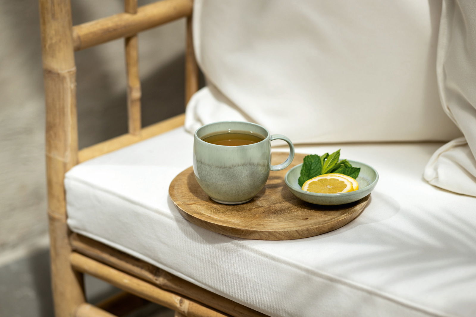 Grüner Becher mit Henkel auf einem Holztablett, neben einem Teller mit Zitronenscheibe und Minzblättern, auf einem weißen Kissenstuhl, seitliche Perspektive