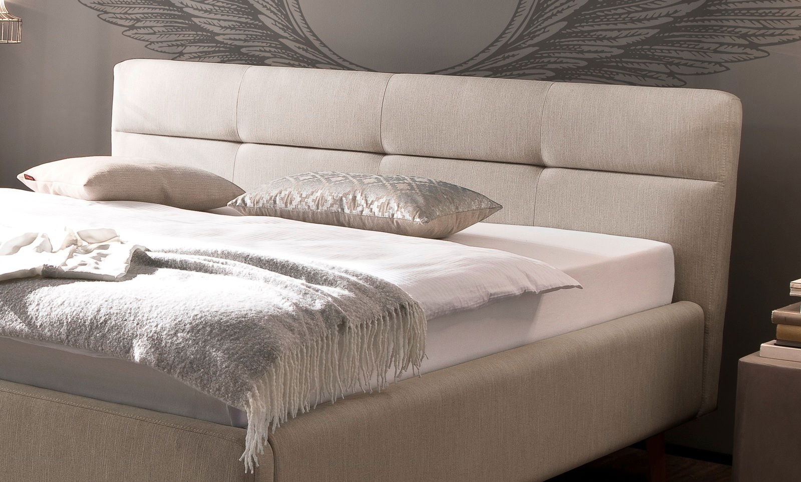 Beiges Polsterbett mit Kopfteil, seitliche Perspektive, dekoriert mit Kissen und Decke.
