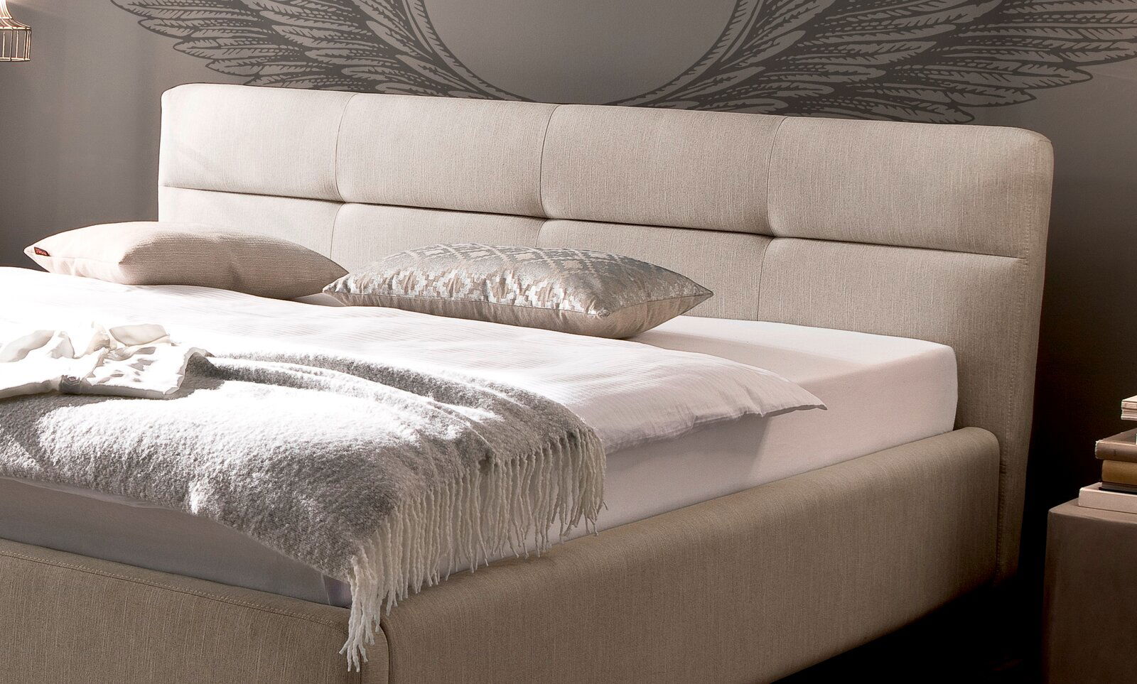 Beiges Polsterbett mit Kopfteil, seitliche Perspektive, dekoriert mit Kissen und Decke.