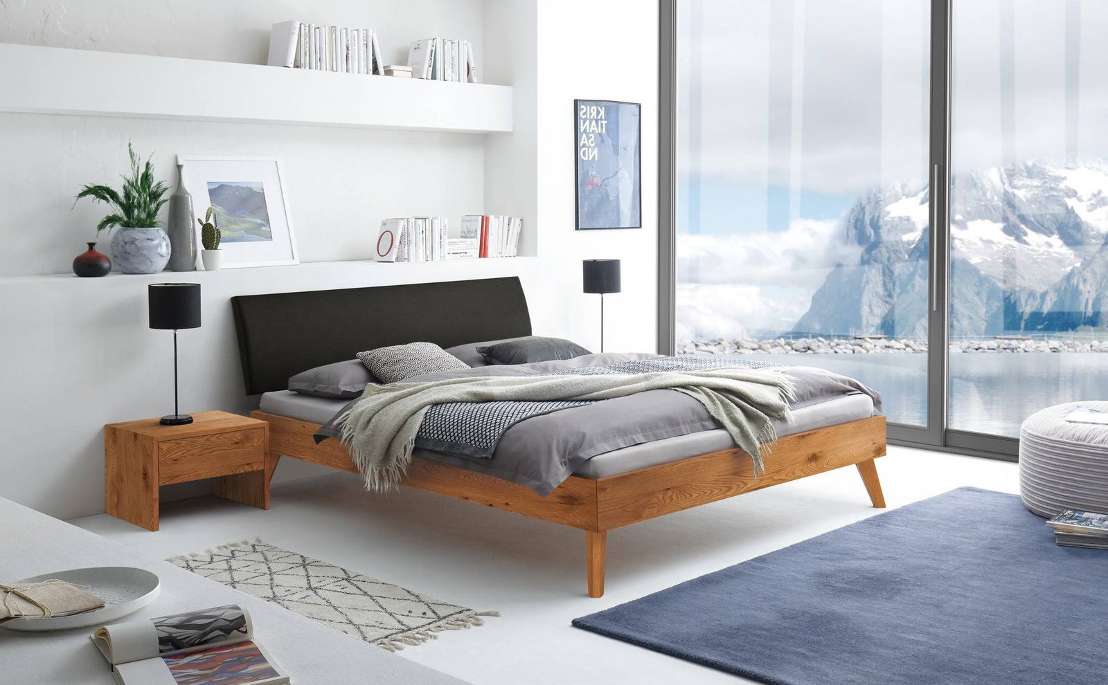 Modernes Bettgestell aus Holz mit schwarzem Kopfteil, seitliche Perspektive, in einem hellen Raum mit großen Fenstern und Bergblick.