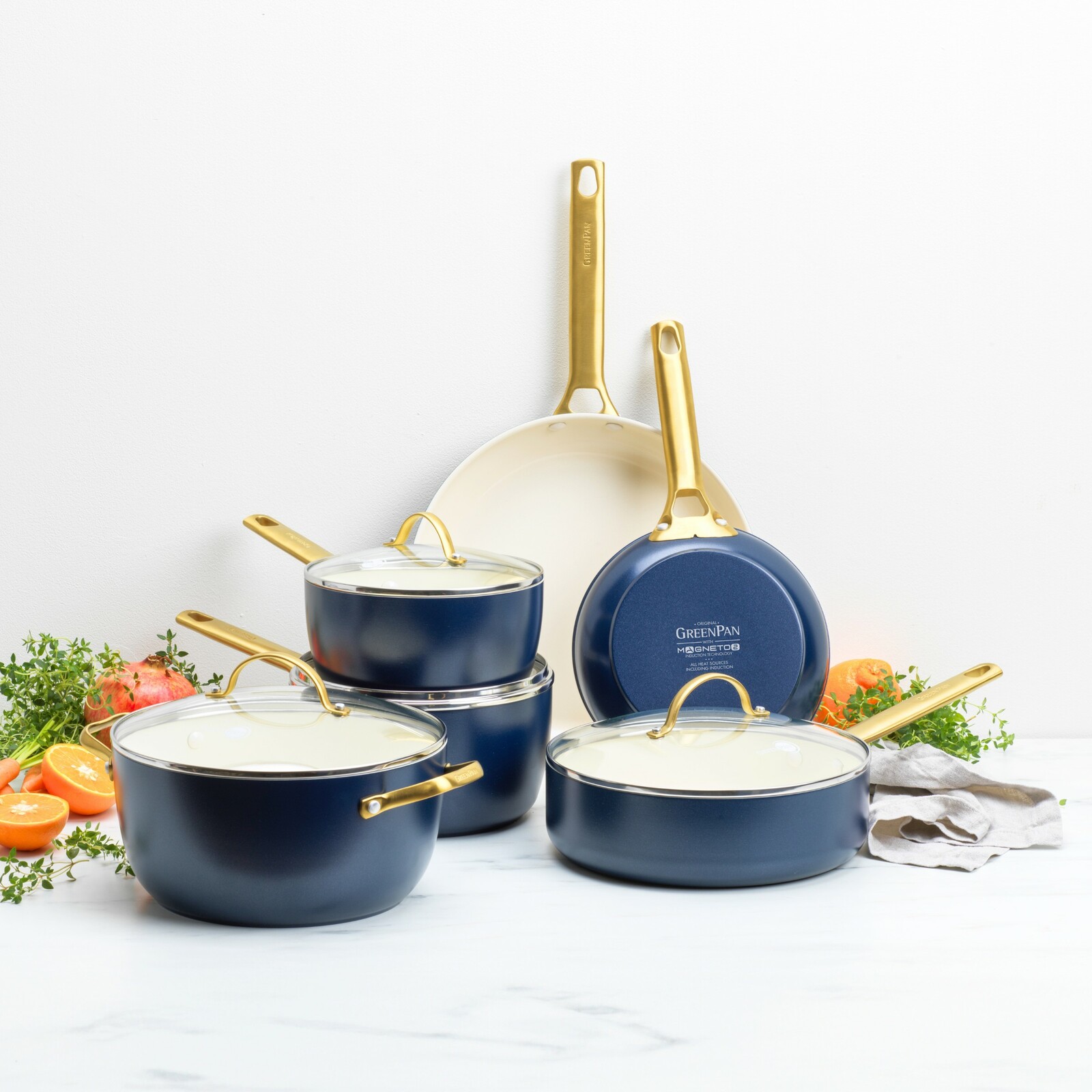 Sechsteiliges Topfset in Blau mit goldenen Griffen, bestehend aus Töpfen und Pfannen, auf einer weißen Oberfläche mit frischen Kräutern und Orangen im Hintergrund, seitliche Perspektive.