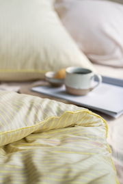 Nahaufnahme einer gestreiften Bettwäsche in sanften Gelbtönen, mit einem Kissen im Hintergrund und einer Tasse auf einem Tablett. Perspektive von der Seite.