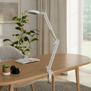 Weiße, verstellbare LED-Schreibtischlampe mit Klemmfuß und Gelenkarm auf einem Holztisch im hellen Wohnzimmer, Seitenansicht schräg von vorn