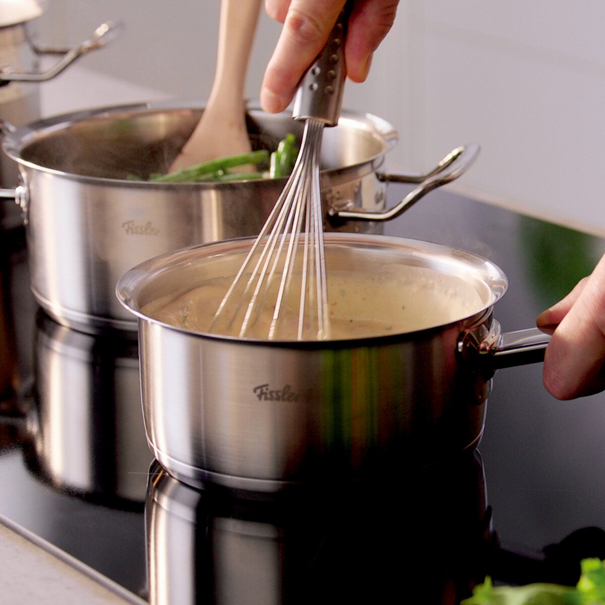 Fissler Topfset 5-tlg. ORIGINAL-PROFI COLLECTION 2 Nahaufnahme eines Topfsets auf einem Herd, in dem eine Person mit einem Schneebesen rührt. Perspektive von der Seite.