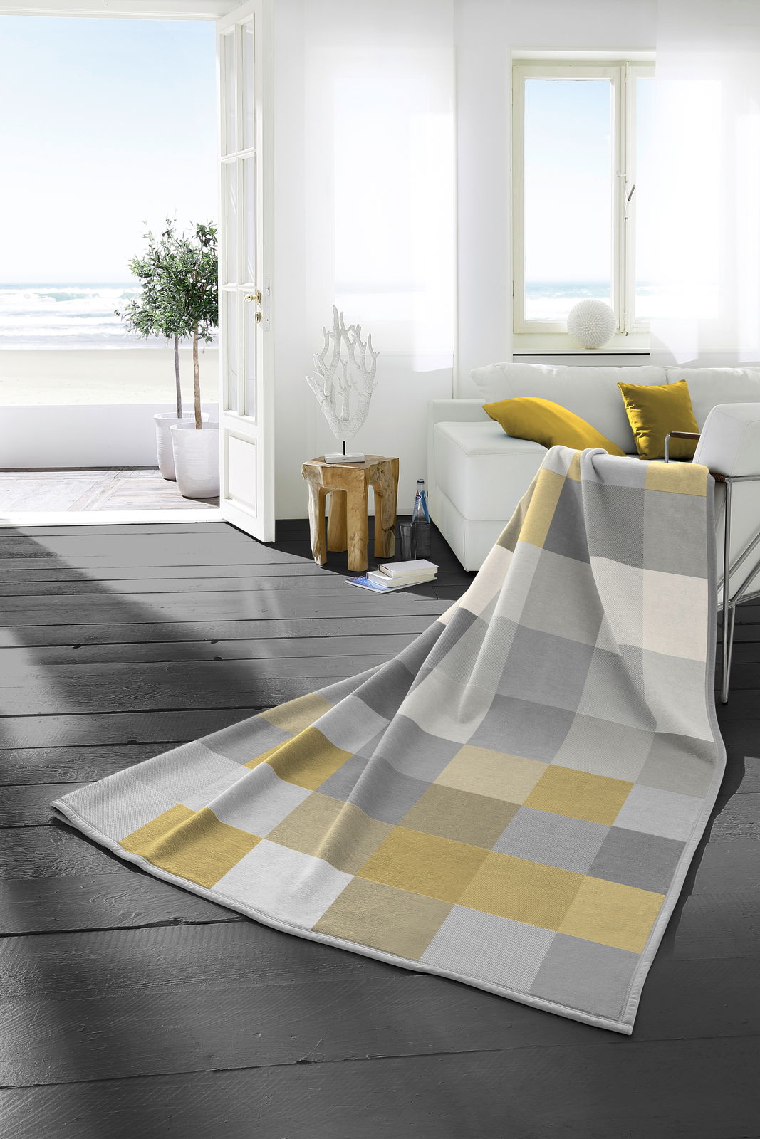 biederlack Heimdecke COTTON HOME Heimdecke 150x200 cm in einem modernen Wohnzimmer mit Blick auf das Meer, in einem karierten Muster aus Grau- und Gelbtönen, drapiert über einem Sofa.
