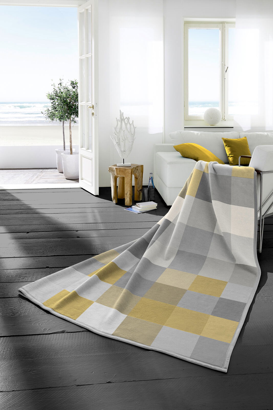 Heimdecke 150x200 cm in einem modernen Wohnzimmer mit Blick auf das Meer, in einem karierten Muster aus Grau- und Gelbtönen, drapiert über einem Sofa.