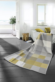 Heimdecke 150x200 cm in einem modernen Wohnzimmer mit Blick auf das Meer, in einem karierten Muster aus Grau- und Gelbtönen, drapiert über einem Sofa.