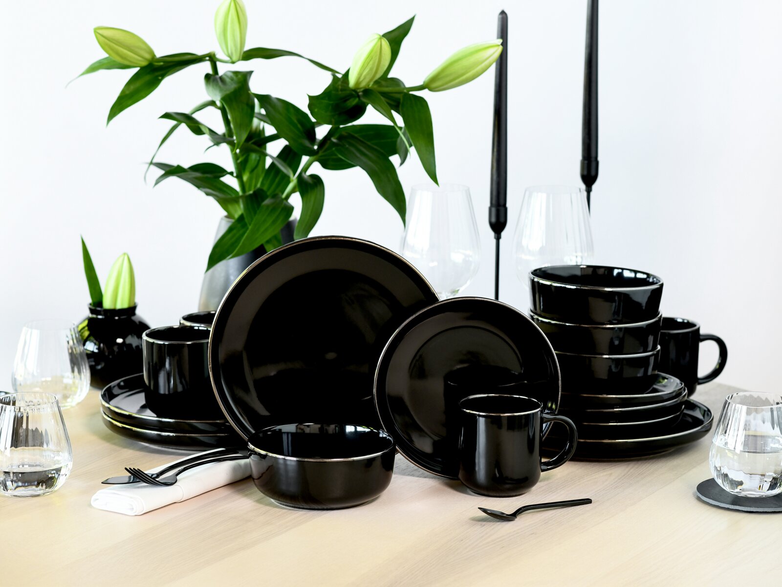 creaTable Kombiservice 16-tlg NORDIC FESTIVE Schwarzes, glänzendes Geschirrset aus Keramik mit Tellern, Schalen und Tassen auf einem gedeckten Holztisch, dekoriert mit Gläsern, Kerzenhaltern und Blumen im Hintergrund, perspektivisch aus leicht schräger Frontansicht aufgenommen.
