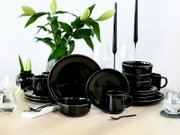 Schwarzes, glänzendes Geschirrset aus Keramik mit Tellern, Schalen und Tassen auf einem gedeckten Holztisch, dekoriert mit Gläsern, Kerzenhaltern und Blumen im Hintergrund, perspektivisch aus leicht schräger Frontansicht aufgenommen.