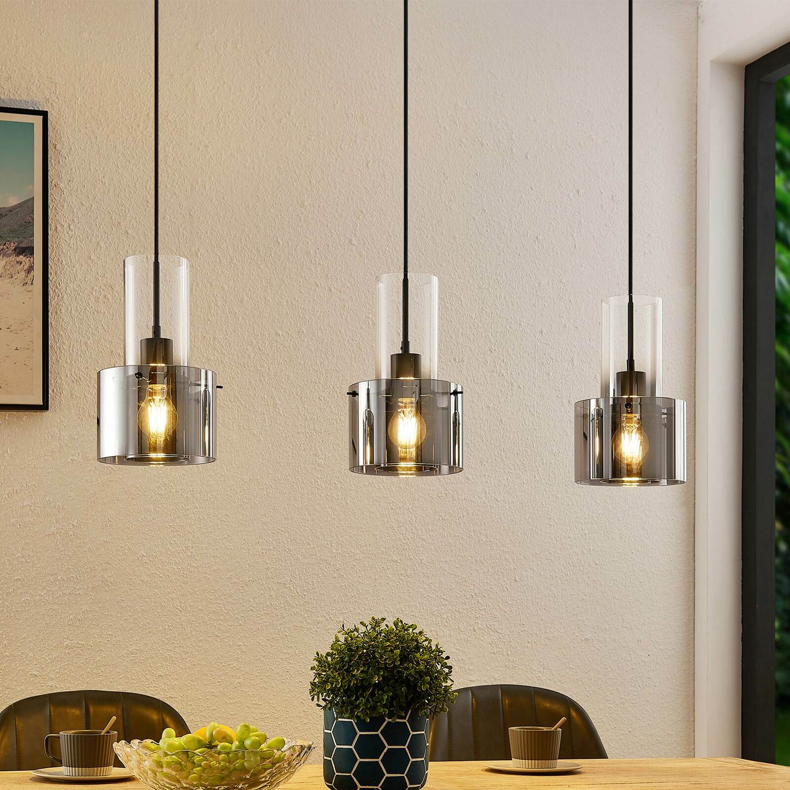 Drei moderne Hängeleuchten mit transparenten Glasschirmen, die von der Decke über einem Esstisch hängen. Perspektive: Frontalansicht.