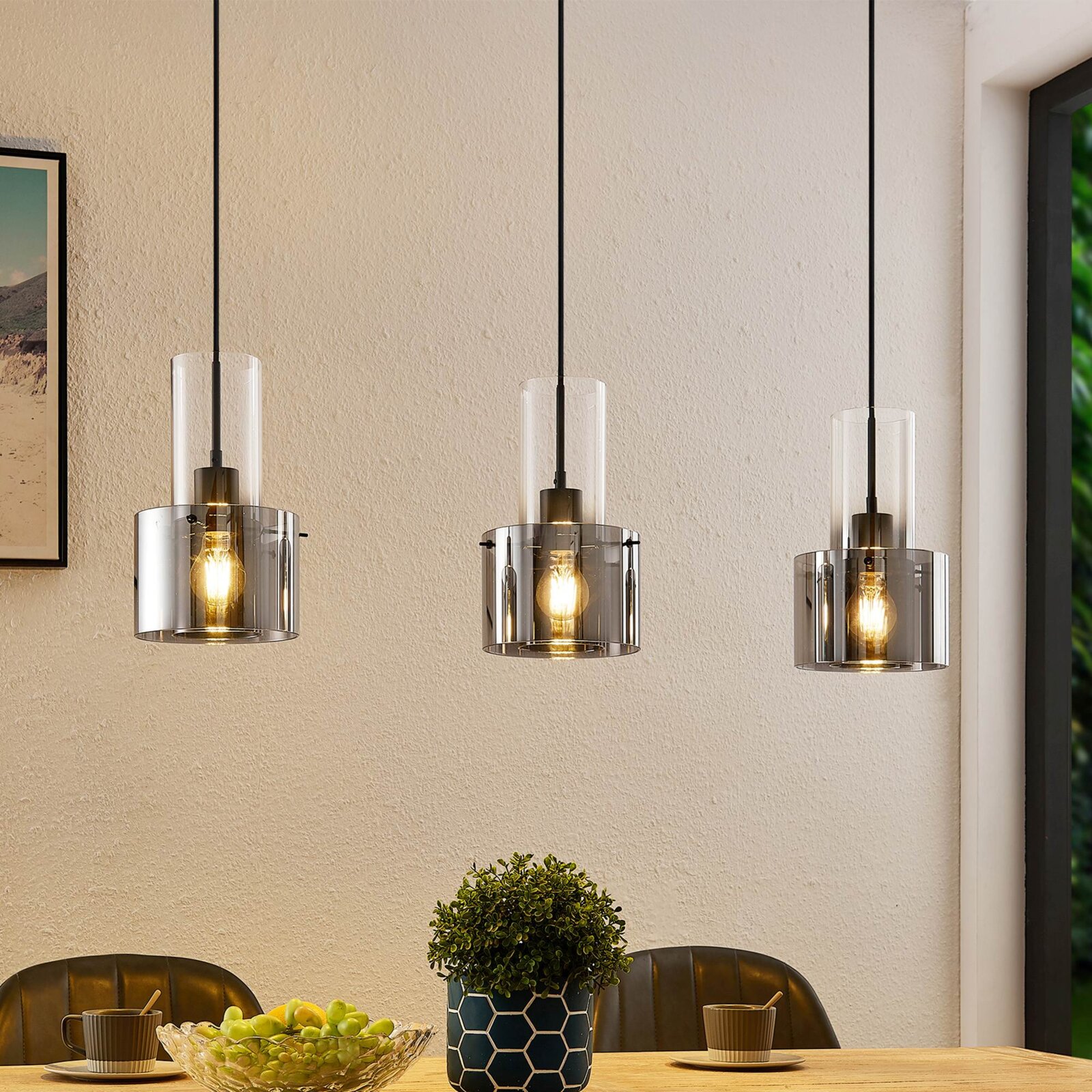 Drie moderne hanglampen met transparante glazen kappen, die vanaf het plafond boven een eettafel hangen. Perspectief: vooraanzicht.
