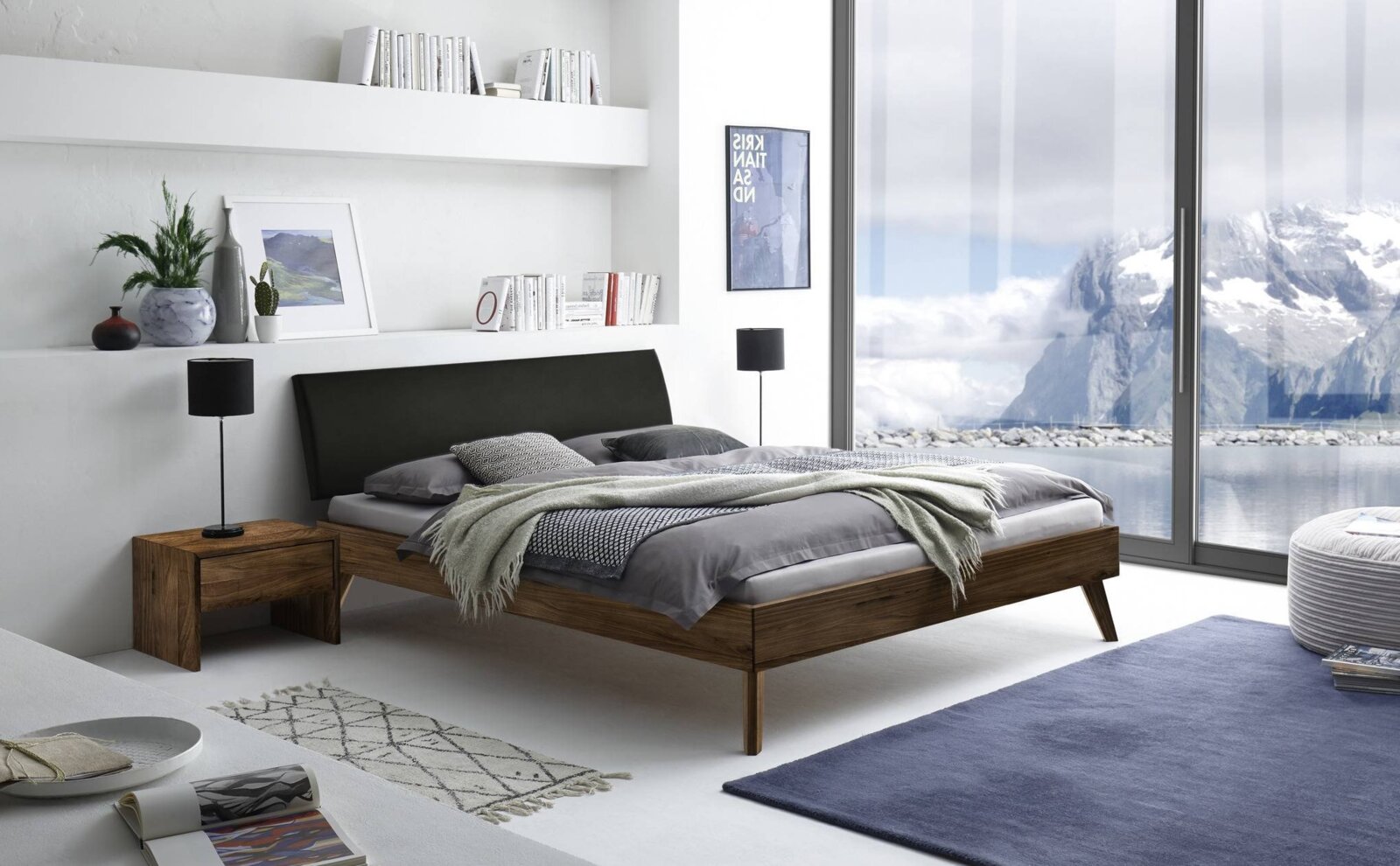 Modern houten bedframe met zwart hoofdeinde, gezien vanaf de zijkant, in een stijlvolle slaapkamer met grote ramen en uitzicht op de bergen.