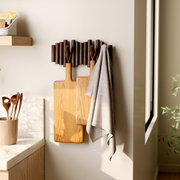 Wandgarderobe mit Holzstäben und Haken, an der zwei Schneidebretter und ein Handtuch hängen, seitliche Perspektive.