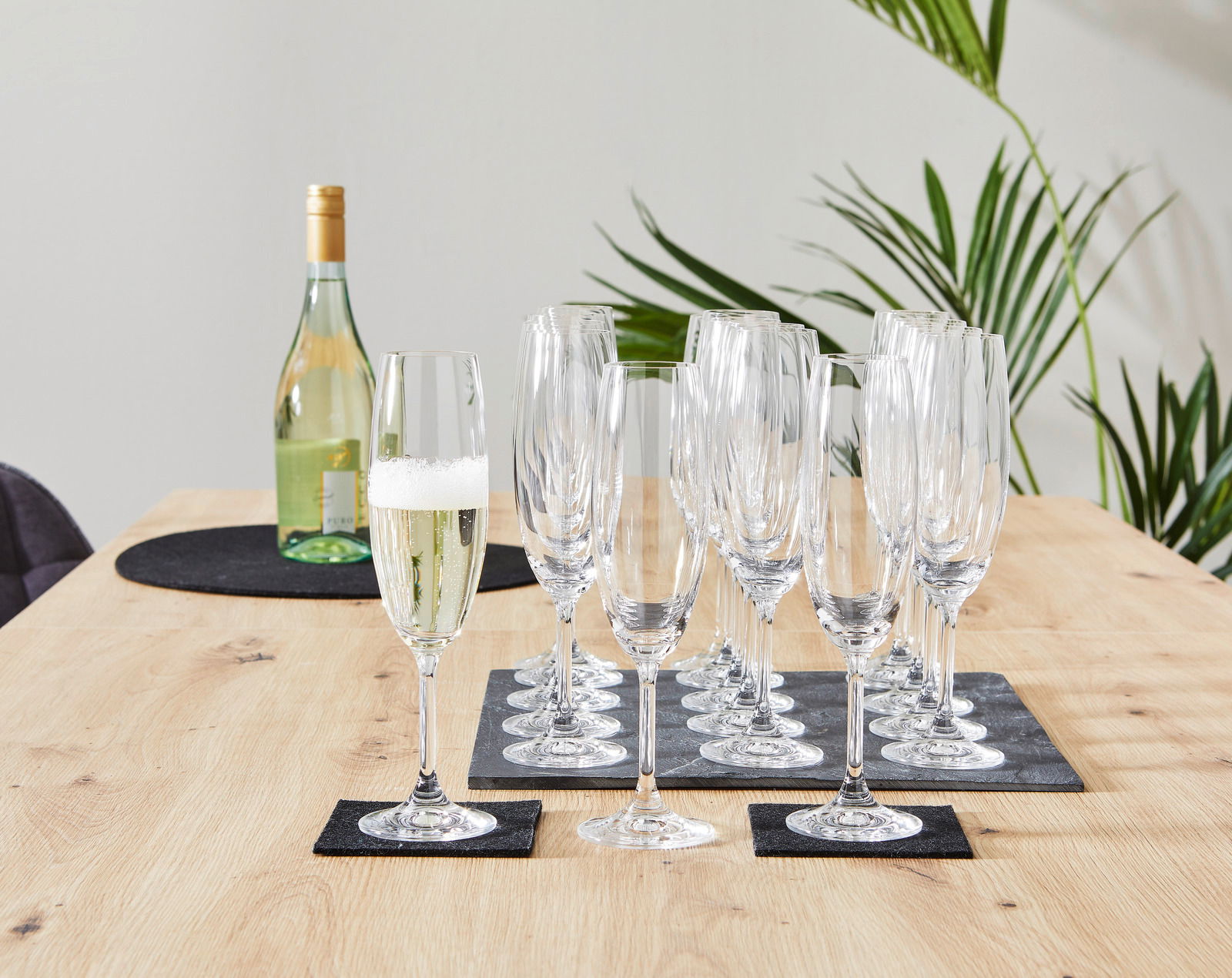 Sektglas-Set mit 18 Gläsern auf einem Holztisch, seitliche Perspektive, mit einer Flasche Sekt im Hintergrund.