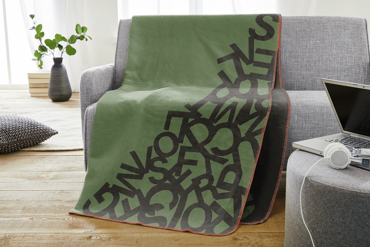 Groene jacquarddeken met een patroon van zwarte letters, gedrapeerd over een grijze bank in een moderne woonkamer.