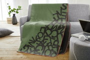 Groene jacquarddeken met een patroon van zwarte letters, gedrapeerd over een grijze bank in een moderne woonkamer.