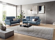 Modernes Wohnzimmer mit zwei blauen Ledersofas, einem runden 3er-Sofa im Vordergrund. Die Perspektive zeigt das Sofa von vorne, umgeben von stilvollen Dekorationen und einem großen Fenster, das viel Licht hereinlässt.