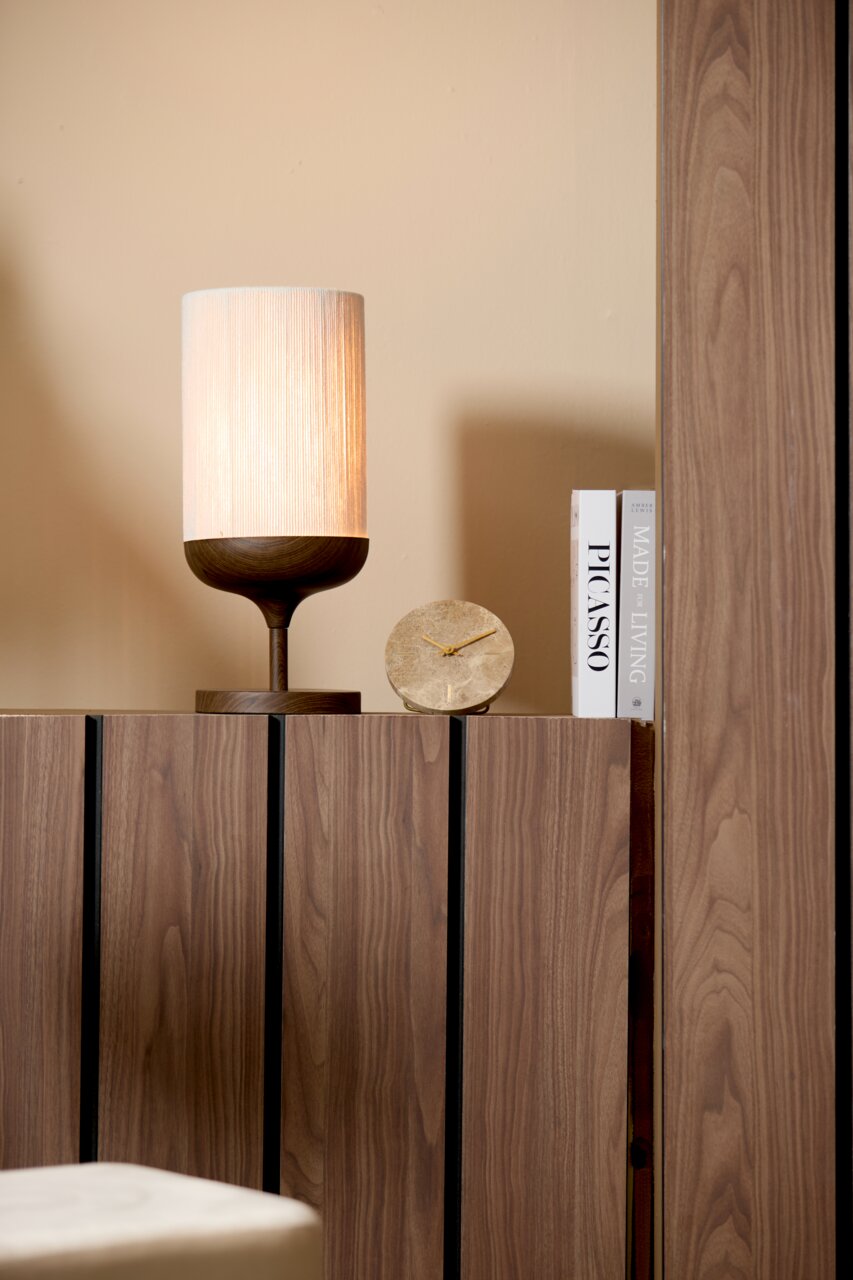Moderne Tischlampe mit zylindrischem, warm leuchtendem Stoffschirm und dunklem Holzfuß auf einem Holzsideboard, daneben runde Stein-Uhr und zwei Bücher Frontansicht leicht von links.