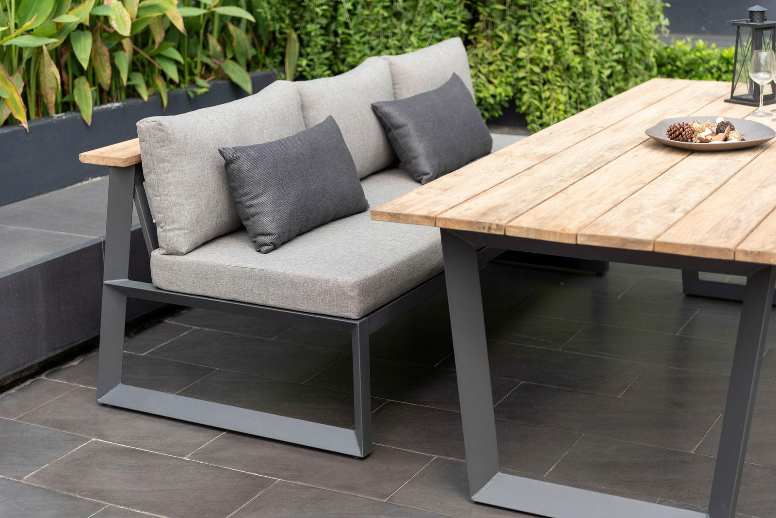 Moderne Sitzbank mit grauen Kissen und Metallrahmen, neben einem Holztisch im Garten, seitliche Perspektive