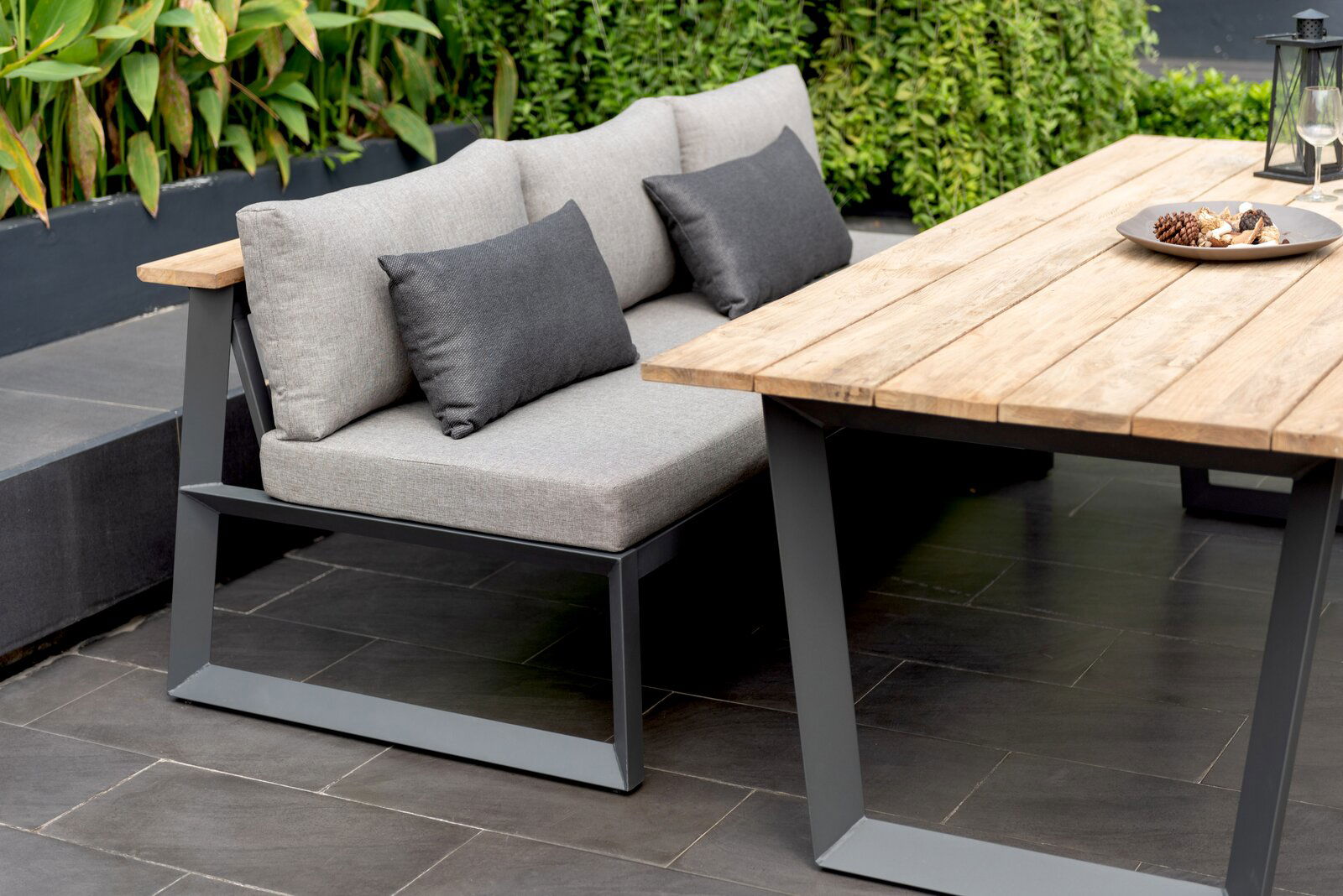 Moderne Sitzbank mit grauen Kissen und Metallrahmen, neben einem Holztisch im Garten, seitliche Perspektive