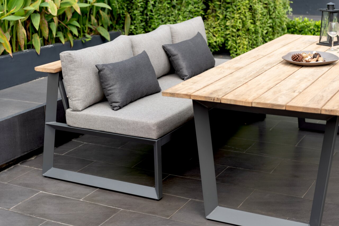 Moderne Sitzbank mit grauen Kissen und Metallrahmen, neben einem Holztisch im Garten, seitliche Perspektive