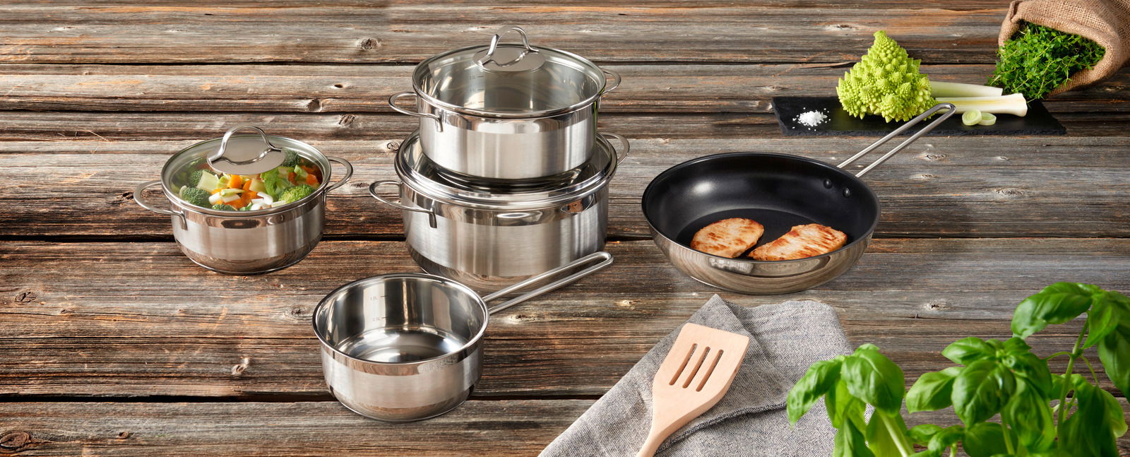Topfset 5-tlg. aus Edelstahl, bestehend aus mehreren Töpfen und einer Pfanne, auf einer rustikalen Holzoberfläche präsentiert. Perspektive von oben.