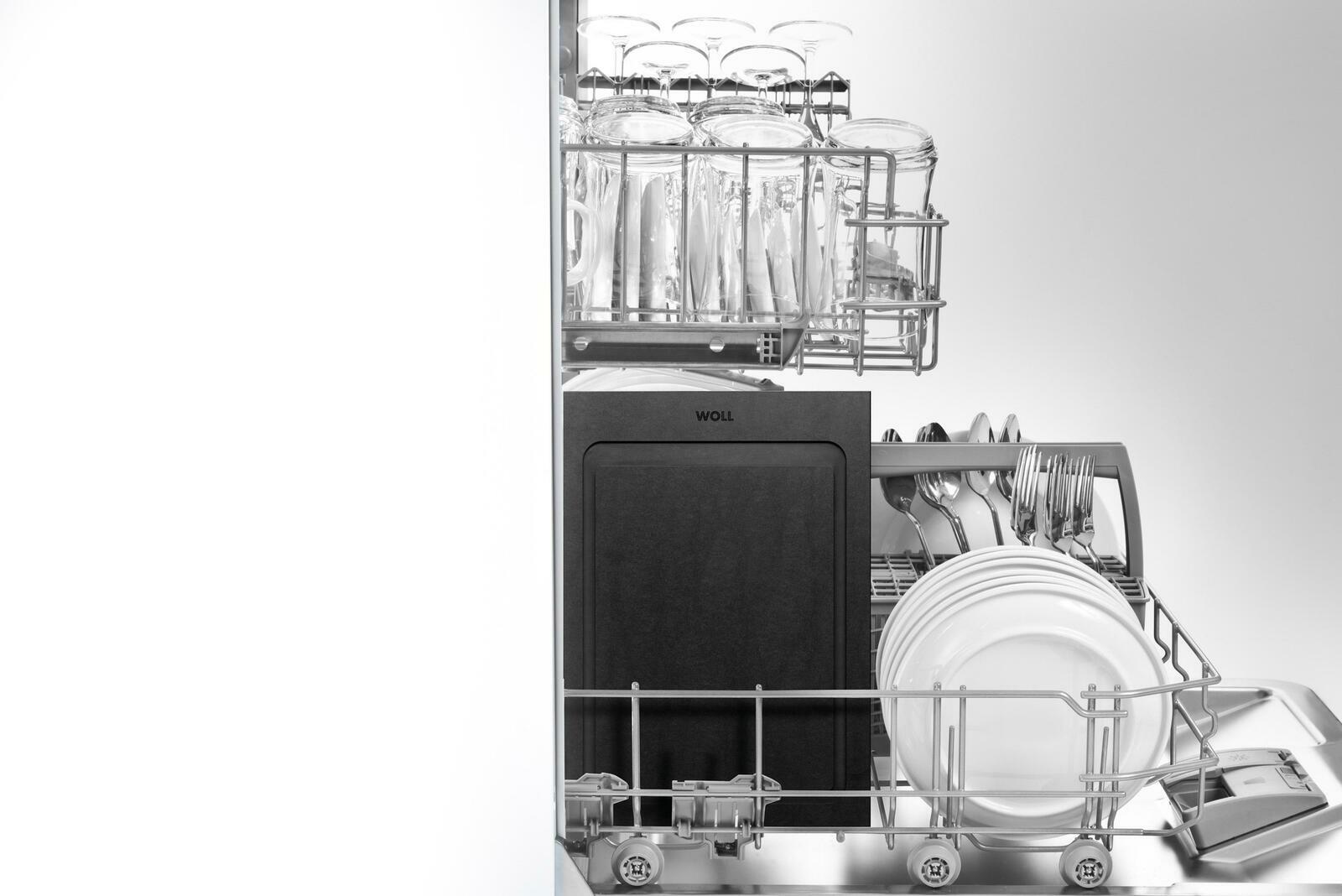 Schwarzes Schneidebrett in einer Spülmaschine, umgeben von Gläsern, Tellern und Besteck, aus der Vorderansicht fotografiert.