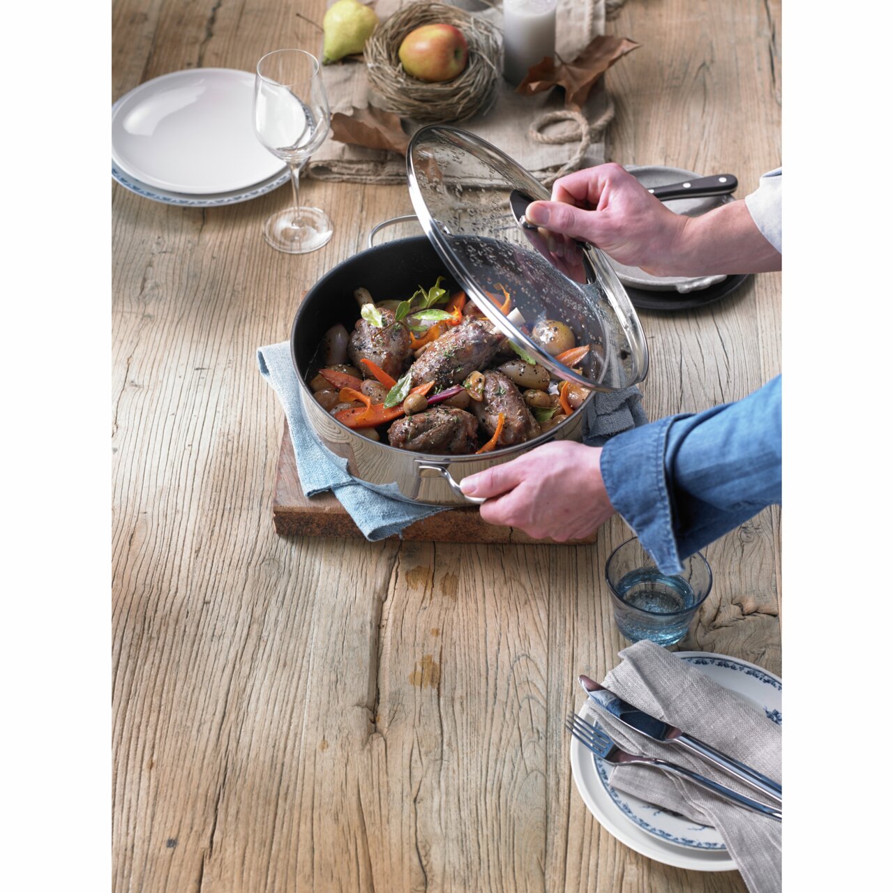 Een serveerpan met glazen deksel, gevuld met gebakken vlees en groenten, gefotografeerd van bovenaf op een houten tafel.