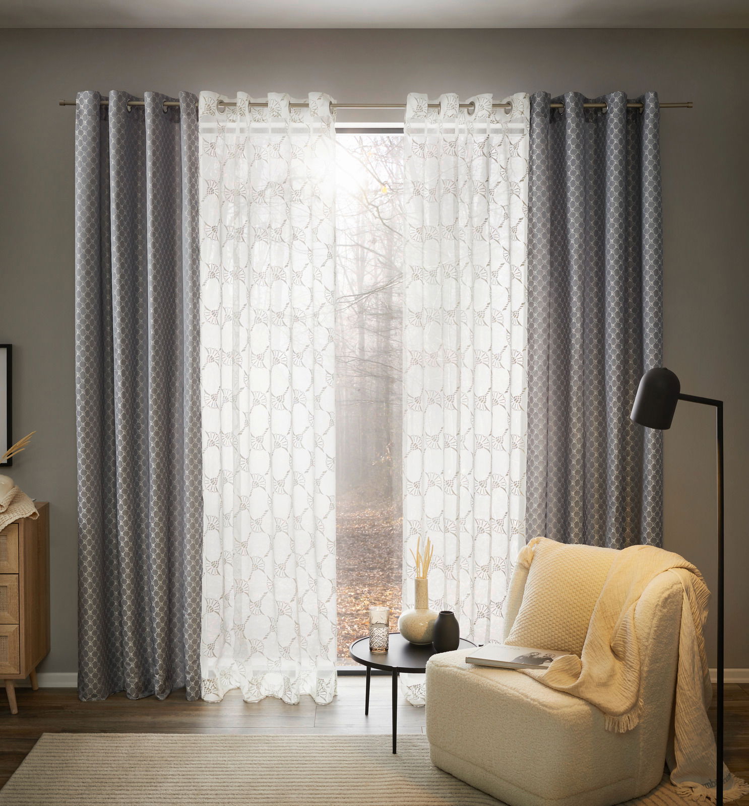 JOOP! Ösenschal J!Allover Silbergrauer Ösenschal in einem gemütlichen Wohnzimmer, frontal betrachtet, mit einem Sessel und einem Beistelltisch im Vordergrund.