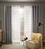 Silbergrauer Ösenschal in einem gemütlichen Wohnzimmer, frontal betrachtet, mit einem Sessel und einem Beistelltisch im Vordergrund.