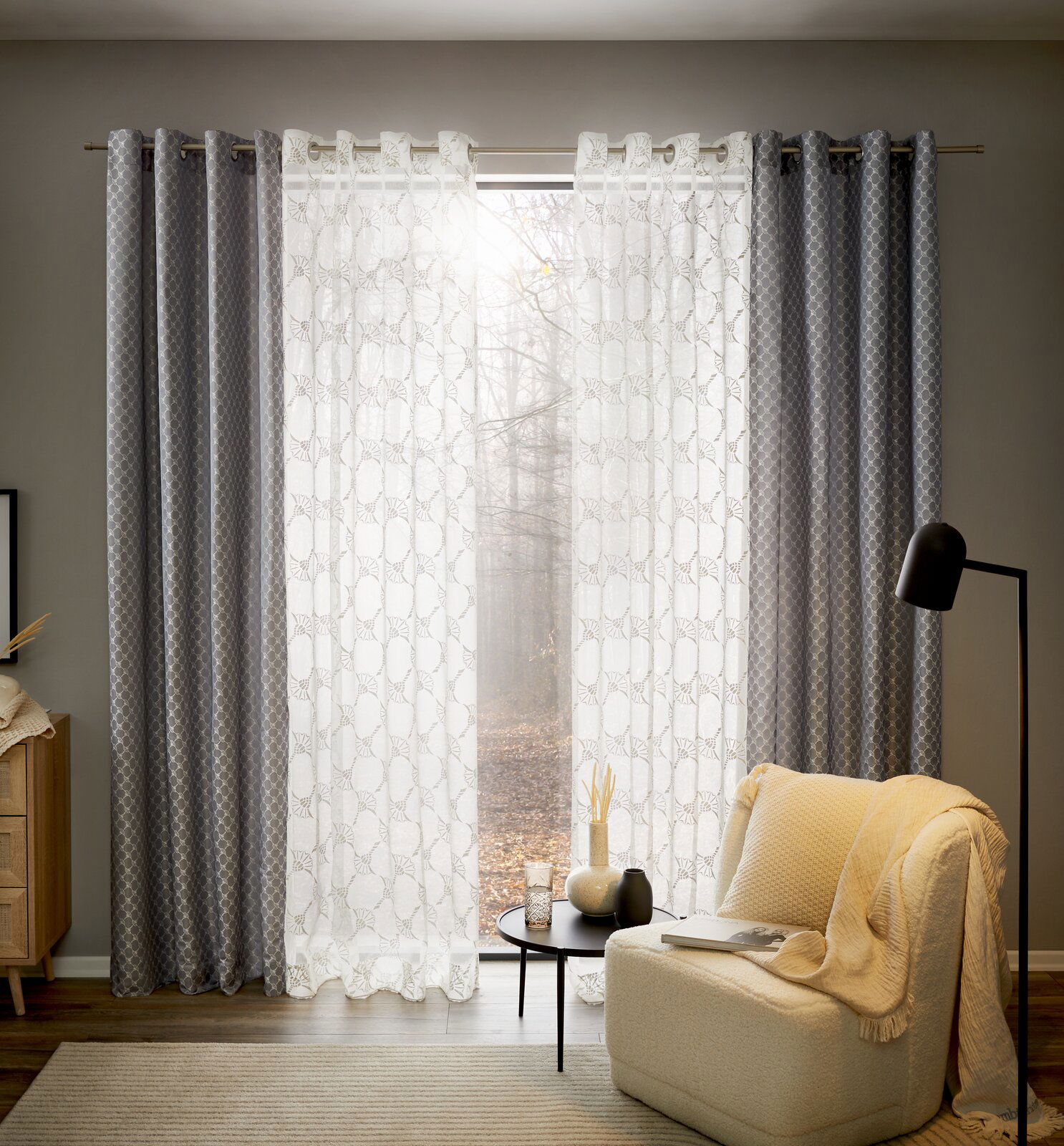 JOOP! Ösenschal J!Allover Silbergrauer Ösenschal in einem gemütlichen Wohnzimmer, frontal betrachtet, mit einem Sessel und einem Beistelltisch im Vordergrund.