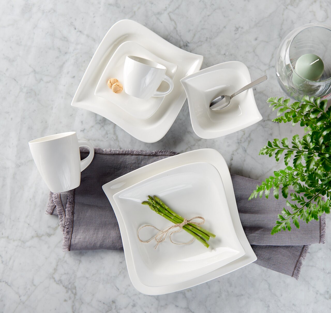 Vogelperspektive auf ein elegantes weißes Kombiservice-Set 'La Musica' mit quadratischen Tellern, Tassen und Schalen auf einem Marmortisch, dekoriert mit Spargel und einem grünen Ei im Glas.